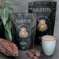 Cacao products including bags, a cup, and cacao beans on a gray surface with a plant.