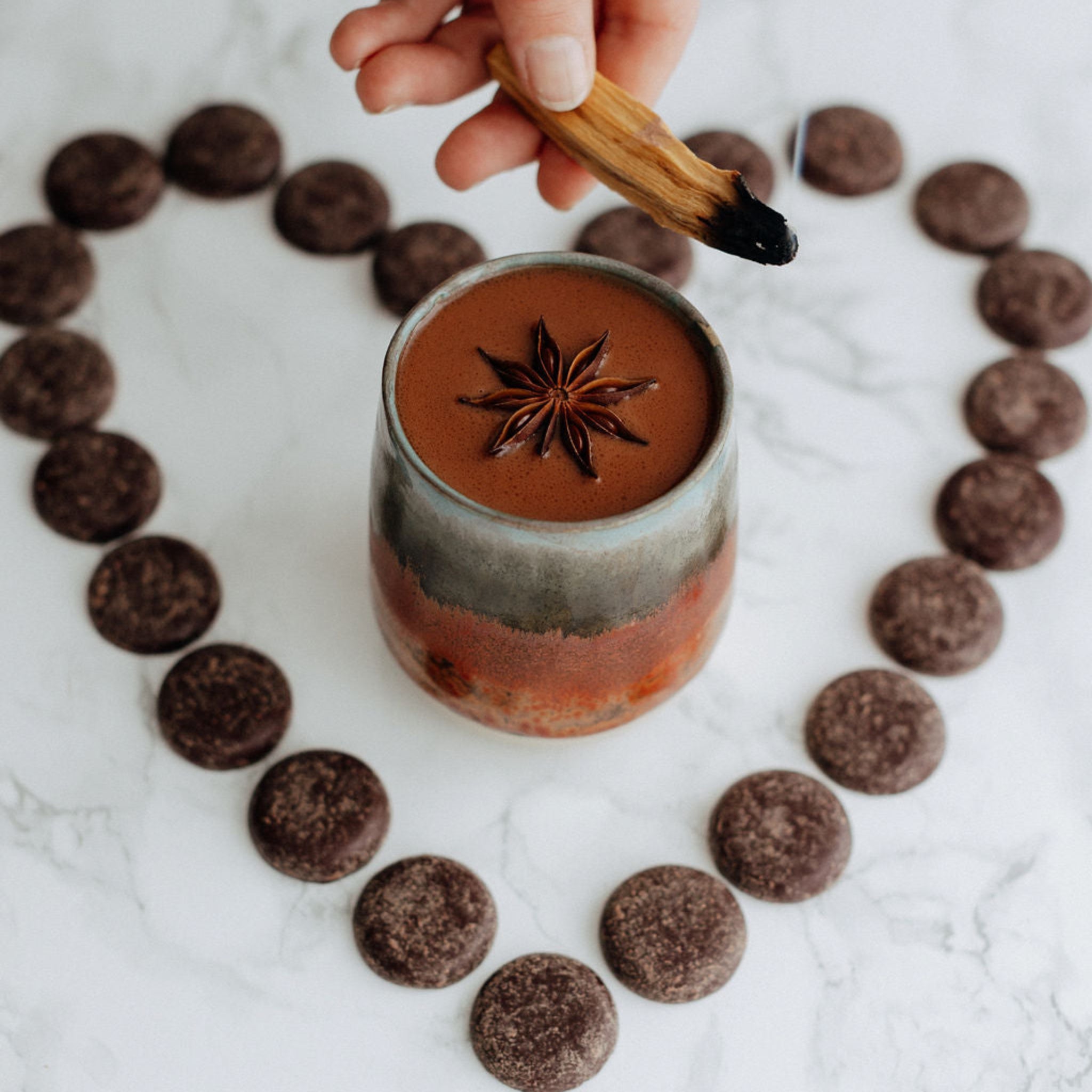 Hand holding palo santo with a star anise over a small cup of ceremonial cacao on a marble surface.