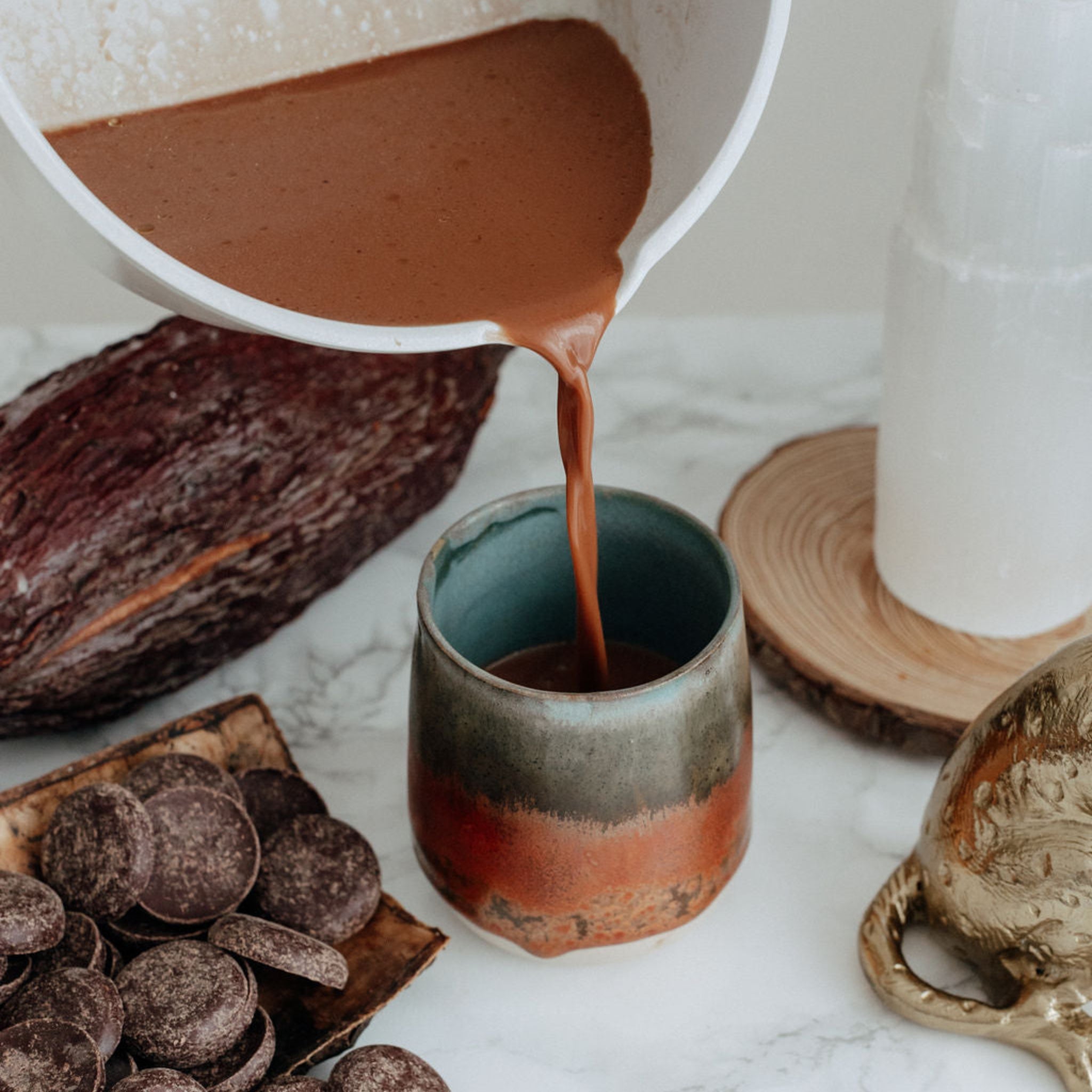 Ceremonial Cacao being poured from a white pot into a ceramic cup on a marble surface with cacao coins around it.