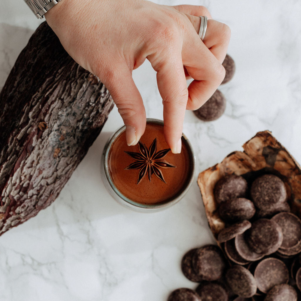 Cup full of ceremonial cacao with female hand placing star anise on top. Placed on marble top surrounded by cacao coins